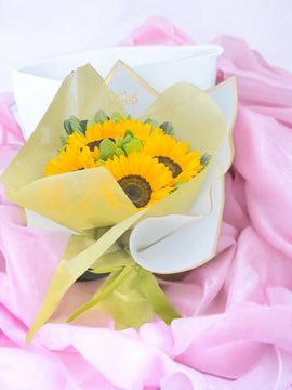 Ramo de girasoles amarillos envueltos en papel amarillo claro, sobre una tela rosa.